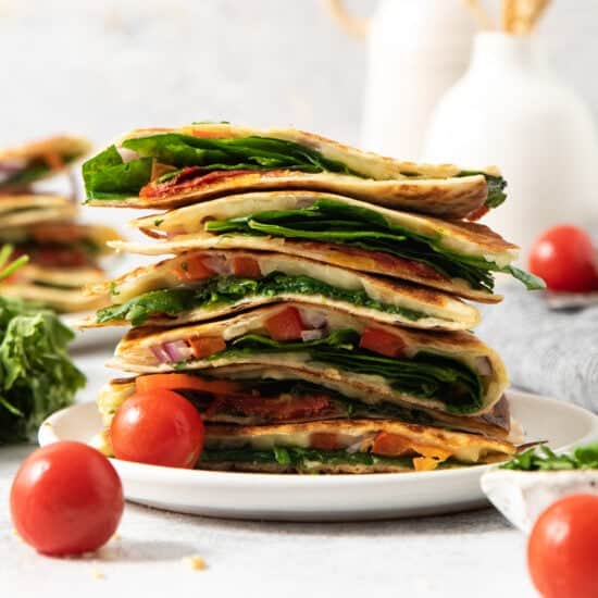 A plate of stacked quesadilla wedges filled with spinach, tomatoes, cheese, and onions. Two cherry tomatoes sit beside the plate.