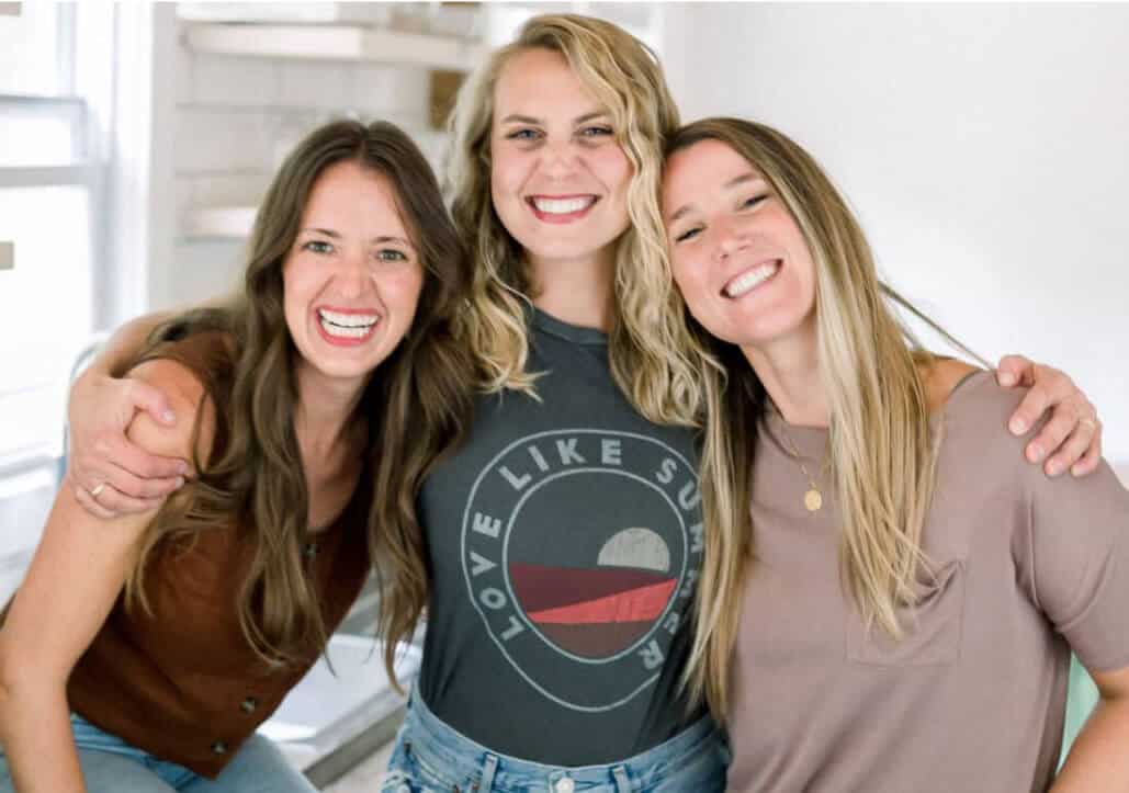 Three women smiling, standing close together with arms around each other in a brightly lit room. The woman in the middle is wearing a graphic t-shirt.