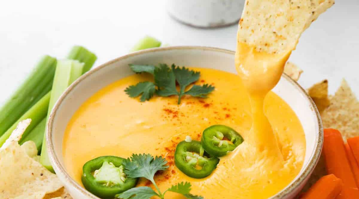 A bowl of cheese dip garnished with sliced jalapeños and cilantro. A tortilla chip is dipped into the cheese. Celery and carrot sticks are placed nearby.