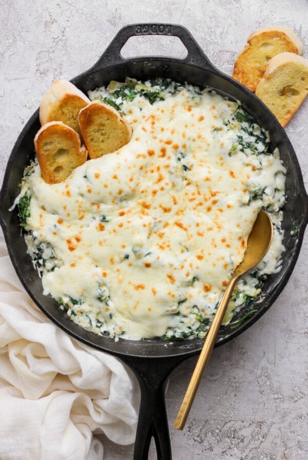 A cast iron skillet filled with baked spinach artichoke dip topped with melted cheese, accompanied by toasted bread slices and a gold spoon.