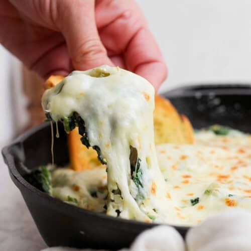 A hand lifts a portion of cheesy spinach dip from a skillet, showing melted cheese stretching. Garlic bread slices are visible in the background.