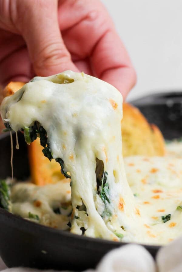 A hand lifts a portion of cheesy spinach dip from a skillet, showing melted cheese stretching. Garlic bread slices are visible in the background.