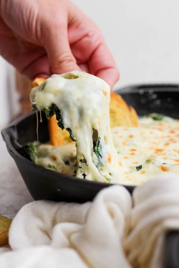 A hand lifts a piece of bread covered in melted cheese and spinach dip from a cast iron skillet.