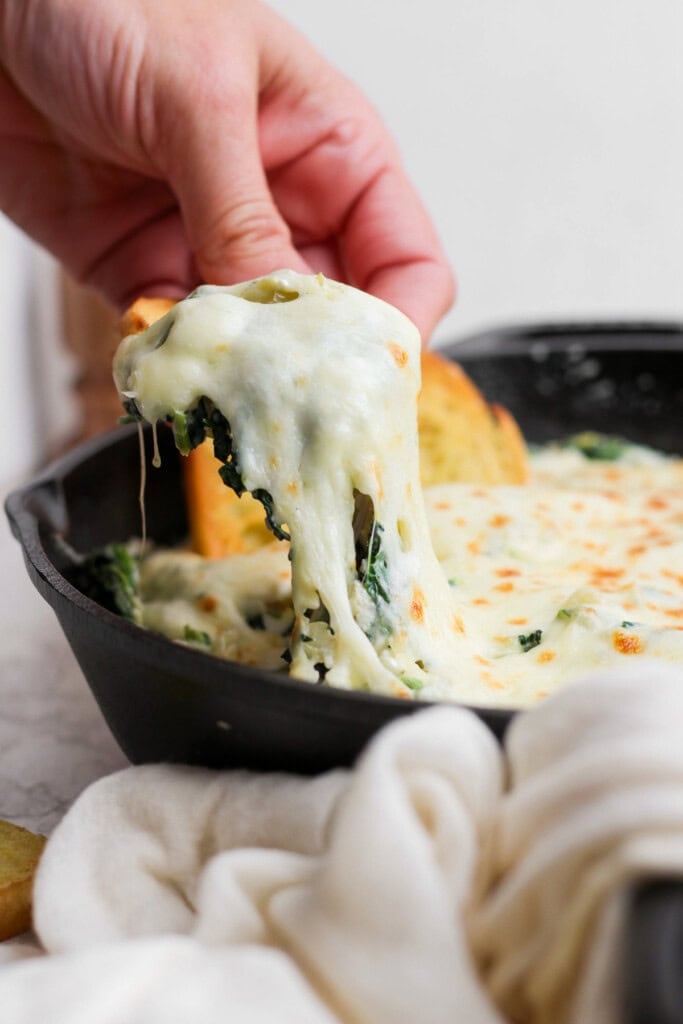 A hand lifts a piece of bread covered in melted cheese and spinach dip from a cast iron skillet.