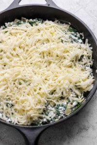A cast iron skillet filled with a mixture of spinach and cheese, topped with a generous layer of shredded cheese, ready to be baked.
