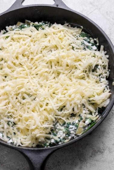A cast iron skillet filled with a mixture of spinach and cheese, topped with a generous layer of shredded cheese, ready to be baked.