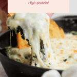 A hand lifts a cheesy, spinach artichoke dip from a skillet, with melted cheese stretching and toast visible in the background.