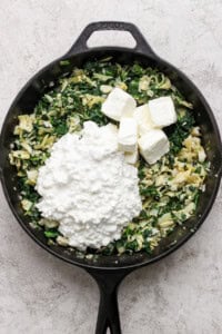 A cast iron skillet filled with chopped spinach, artichokes, and onions, topped with cubes of cream cheese and a heap of cottage cheese.