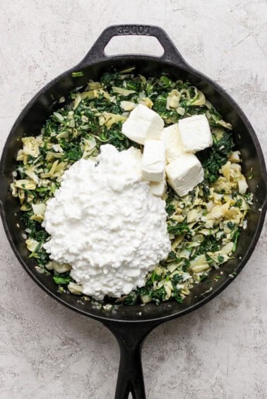 A cast iron skillet filled with chopped spinach, artichokes, and onions, topped with cubes of cream cheese and a heap of cottage cheese.