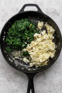 Chopped spinach, artichoke hearts, and sautéed onions in a black cast iron skillet on a light countertop.