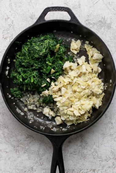 Chopped spinach, artichoke hearts, and sautéed onions in a black cast iron skillet on a light countertop.