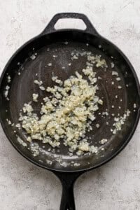 Diced onions and minced garlic being sautéed in a black cast iron skillet on a light-colored surface.
