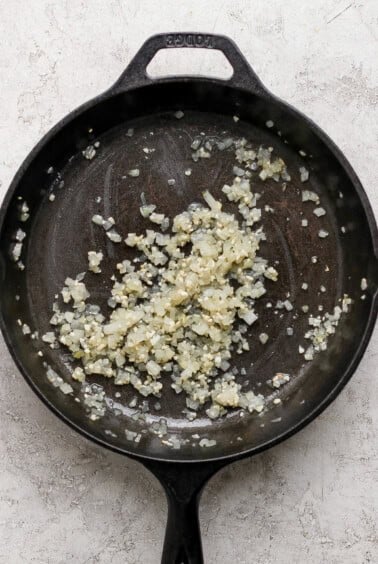 Diced onions and minced garlic being sautéed in a black cast iron skillet on a light-colored surface.