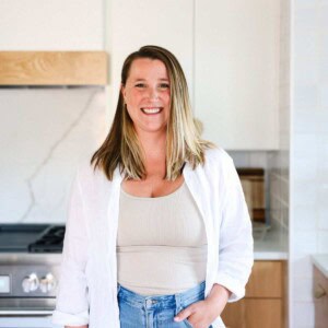 A woman with straight, shoulder-length hair stands smiling in a modern kitchen, wearing a white shirt over a beige top and blue jeans—clearly excited about the cheese knees recipe she’s just prepared.
