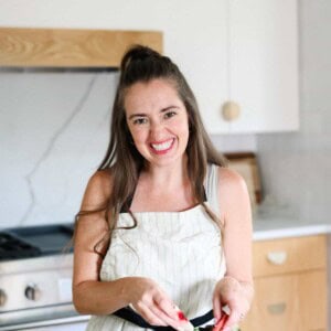 A woman wearing an apron stands in a kitchen, smiling at the camera while preparing food—perhaps about the cheese knees, she's ready to add a playful twist to her recipe.