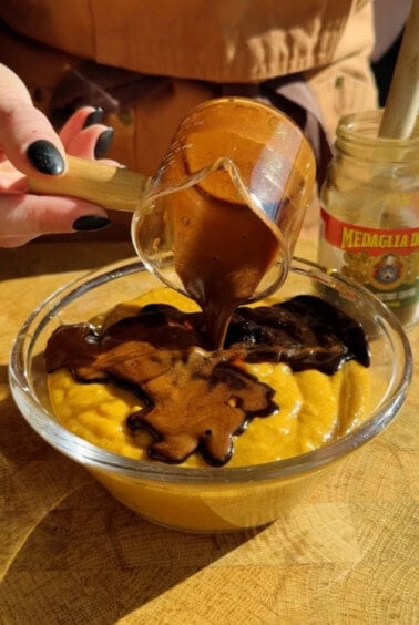 A hand pours thick chocolate sauce from a glass cup onto a bowl of yellow pudding. A jar labeled "Medaglia D’Oro" is visible in the background.