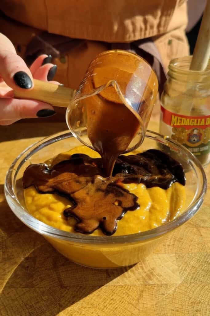 A hand pours thick chocolate sauce from a glass cup onto a bowl of yellow pudding. A jar labeled "Medaglia D’Oro" is visible in the background.