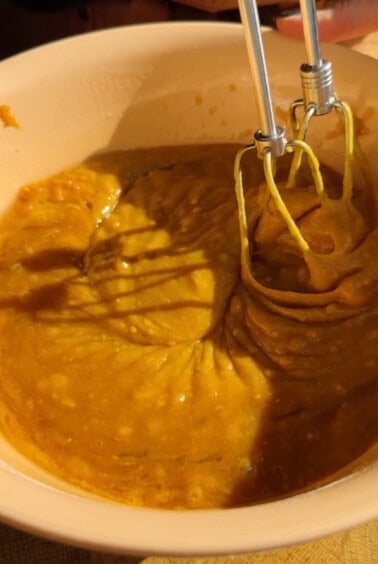 A close-up of a mixing bowl containing thick orange batter being mixed with an electric hand mixer.