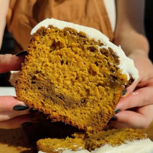 A person holds a thick slice of moist, orange-brown cake with white frosting on top, showing its crumb texture.