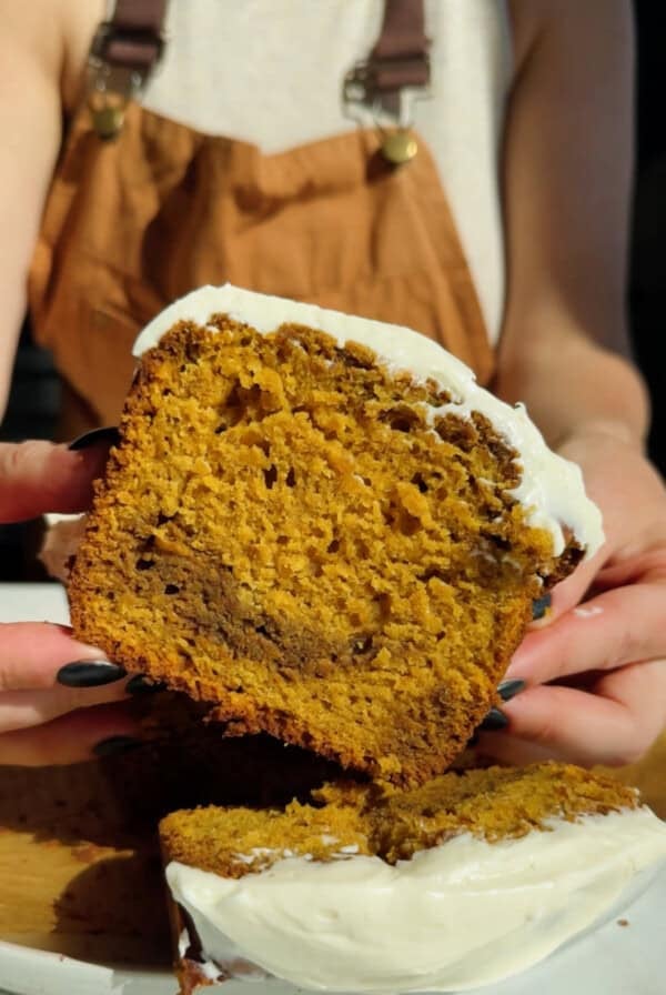 A person holding a thick slice of pumpkin bread with cream cheese frosting, showing the moist texture and dark swirls inside.