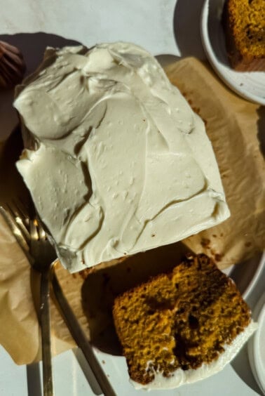 A rectangular cake with white frosting, partially sliced, sits on parchment paper with a fork beside it; a slice is served on a nearby plate.