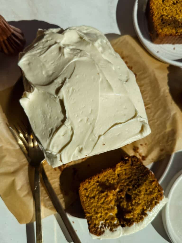 A rectangular cake with white frosting, partially sliced, sits on parchment paper with a fork beside it; a slice is served on a nearby plate.