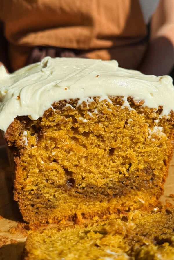 A sliced loaf of pumpkin bread with cream cheese frosting on top sits on a wooden tray, with a fork and a person in the background.