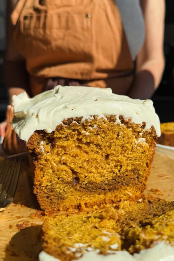 A close-up of a sliced loaf cake with white frosting, sitting on a wooden cutting board; a person in a tan apron stands in the background.
