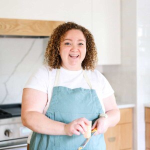 A person with curly hair wearing a white shirt and a blue apron stands in a kitchen, smiling while holding a kitchen tool—ready to share all about the cheese knees.