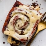 A close-up of a cinnamon roll topped with white icing and chocolate shavings on a plate next to a gold fork.