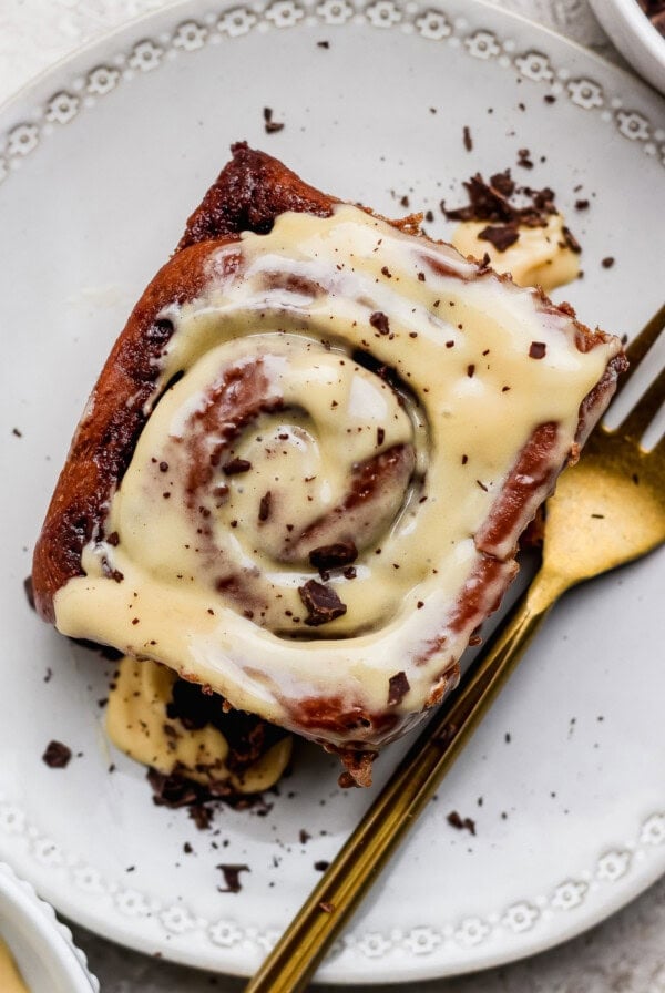 A cinnamon roll topped with cream cheese icing and chocolate shavings on a white plate with a gold fork.