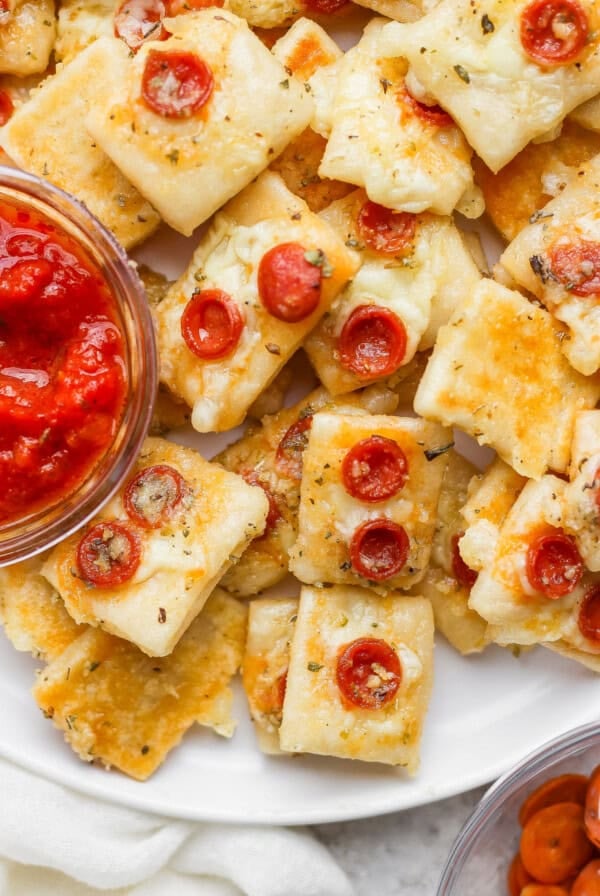 A plate of square mini pizza bites topped with cheese and small pepperoni slices, served with a bowl of marinara sauce for dipping.