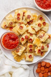 A plate of rectangular cheesy bread bites topped with mini pepperoni slices, served with bowls of marinara sauce and extra mini pepperoni on the side.