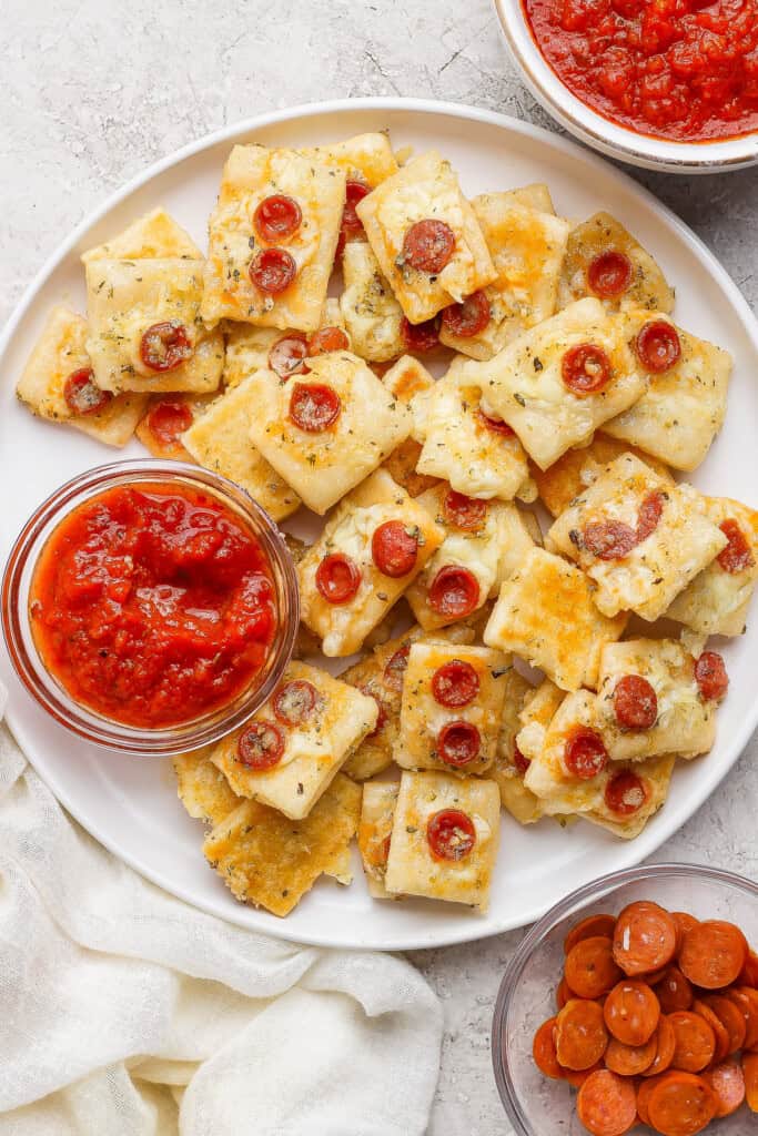 A plate of rectangular cheesy bread bites topped with mini pepperoni slices, served with bowls of marinara sauce and extra mini pepperoni on the side.