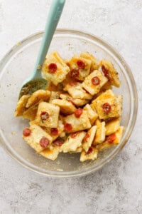 A glass bowl filled with seasoned crackers, shredded cheese, and mini pepperoni slices, mixed with a light blue spatula.