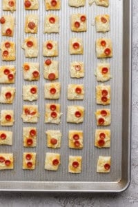 A baking sheet with rows of square puff pastry bites topped with melted cheese and mini pepperoni slices.