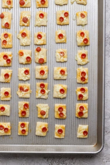 A baking sheet with rows of square puff pastry bites topped with melted cheese and mini pepperoni slices.