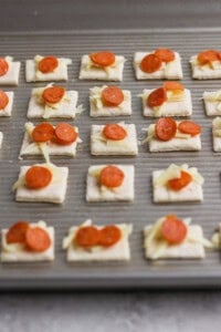A baking sheet with rows of square crackers topped with shredded cheese and small pepperoni slices, ready to be baked.