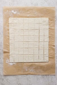 Square pieces of dough are evenly cut and arranged on a sheet of parchment paper atop a light, textured surface.