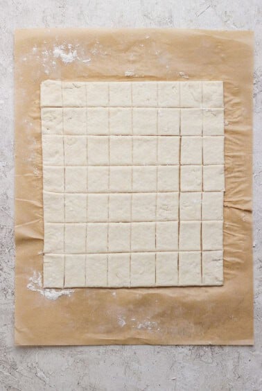 Square pieces of dough are evenly cut and arranged on a sheet of parchment paper atop a light, textured surface.