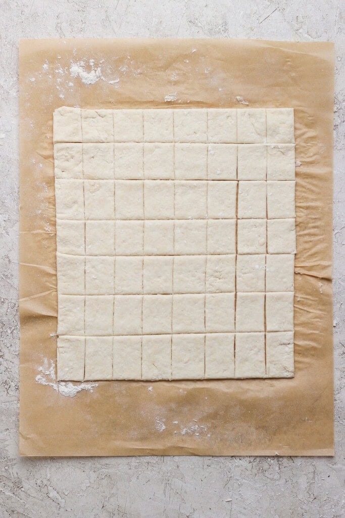 Square pieces of dough are evenly cut and arranged on a sheet of parchment paper atop a light, textured surface.