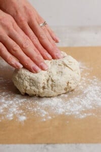 Two hands pressing down on a ball of dough on a floured surface, preparing to knead or shape it.