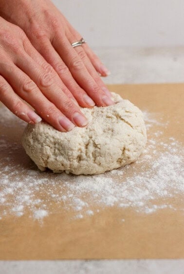 Two hands pressing down on a ball of dough on a floured surface, preparing to knead or shape it.