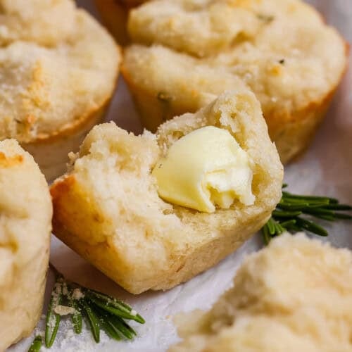 A close-up of a biscuit broken open with a pat of butter melting inside, garnished with fresh rosemary sprigs.