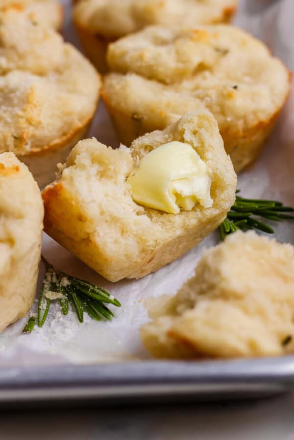 A close-up of a biscuit cut open with a pat of butter melting inside, surrounded by other biscuits and sprigs of rosemary on a tray.