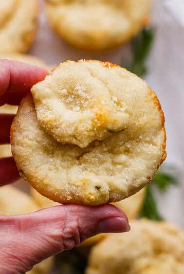 A hand holding a round, baked biscuit topped with grated cheese, with more biscuits blurred in the background.