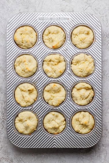 A muffin tin containing twelve baked muffins with a light golden top, set on a textured white surface.
