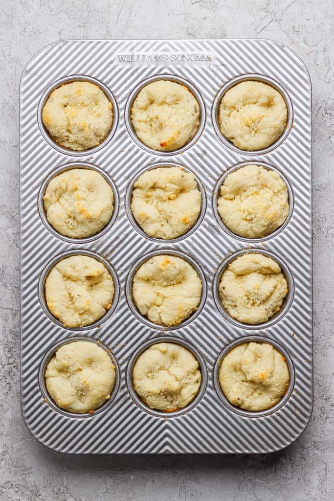 A muffin tin containing twelve baked muffins with a light golden top, set on a textured white surface.