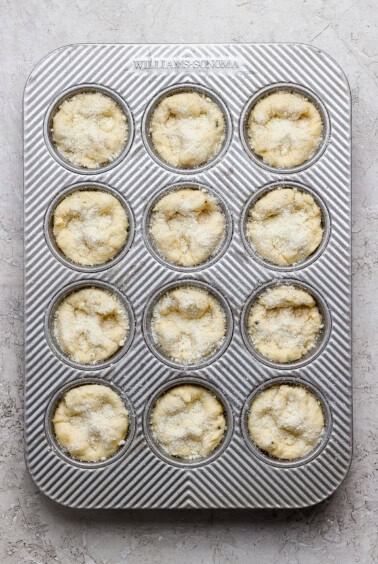 A metal muffin tin with twelve unbaked muffin batter portions, sprinkled with sugar, placed on a light textured surface.
