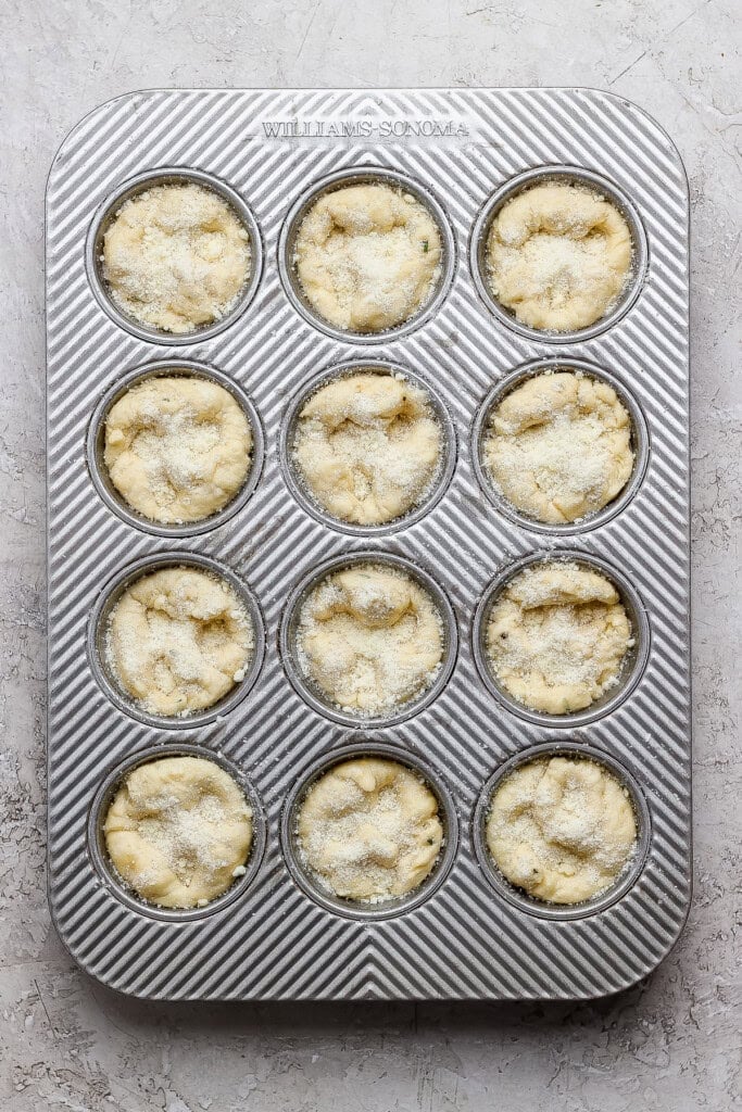 A metal muffin tin with twelve unbaked muffin batter portions, sprinkled with sugar, placed on a light textured surface.